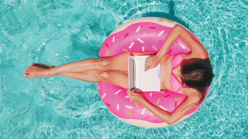 穿着泳衣拿着笔记本电脑的女人坐在粉红色的圆浮在游泳池里